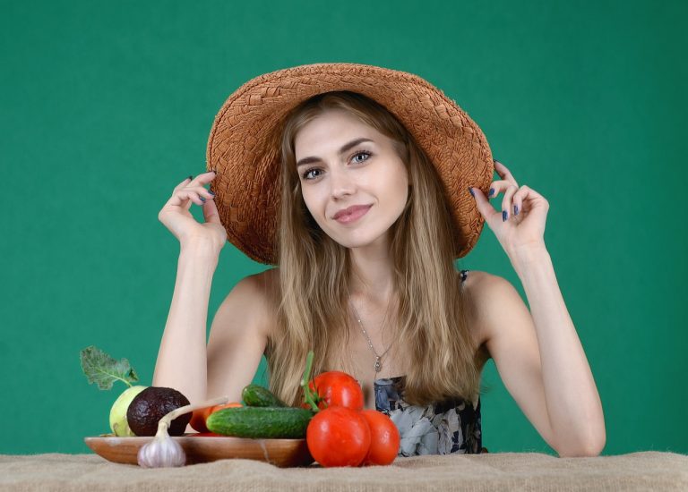 Portrait of a smiling woman wearing a straw hat, seated at a table with fresh vegetables including tomatoes, cucumbers, garlic, and avocado, against a green background.