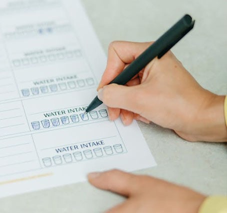 expert A person is using a pen to check off boxes on a printed water intake tracker. The sheet, designed perfectly for a veganiac lifestyle, has rows labeled "WATER INTAKE," with empty boxes ready to be filled as hydration goals are met.