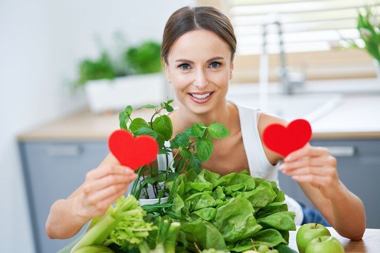 Smiling woman holding two red heart-shaped cutouts in front of a table filled with fresh green vegetables, herbs, and apples, symbolizing healthy eating and love for nutritious food.