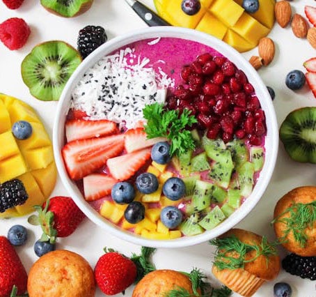 lifestyle A vibrant smoothie bowl topped with sliced strawberries, kiwi, mango, pomegranate seeds, blueberries, coconut flakes, and chia seeds. Perfect for any veganiac, it’s surrounded by fresh fruits and muffins on a white background.
