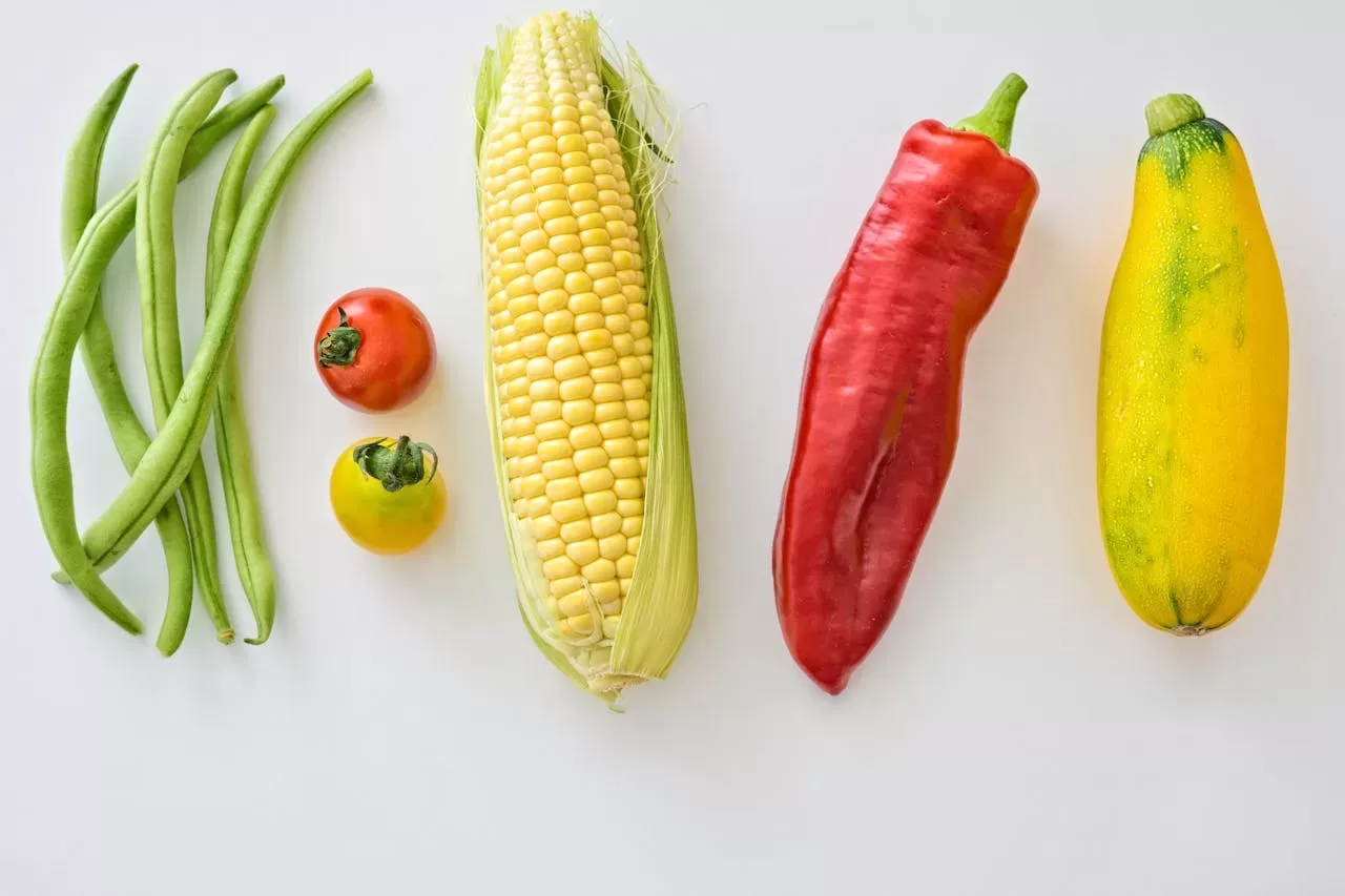 A vibrant vegan display features a row of fresh vegetables and fruit on a white surface, including green beans, two tomatoes (red and yellow), an ear of corn, a red pepper, and a yellow zucchini.
