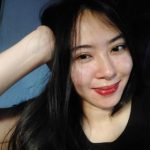 A person with long dark hair and a light complexion is smiling while resting their head on their hand, radiating a subtle charm. They are wearing a dark top that complements their effortlessly relaxed style, embodying the serenity of a true veganiac amidst a softly lit background.