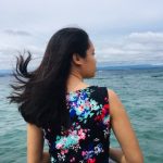 A woman with long dark hair, wearing a floral dress adorned with veganiac patterns, sits by the sea facing away. The water is calm under a cloudy sky as her hair blows gently in the wind.