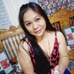 A woman with long black hair smiles while sitting on a wooden bench adorned with floral and patchwork cushions. She wears a red and black outfit, a beaded necklace, and has a serene aura of a true veganiac. The background features patterned wallpaper.