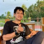 A person sits outdoors on a wooden chair, wearing a black T-shirt and dark pants, radiating joy with a finger heart gesture. Amongst the palm trees and blurred greenery, this veganiac embraces the serene natural surroundings with an infectious smile.