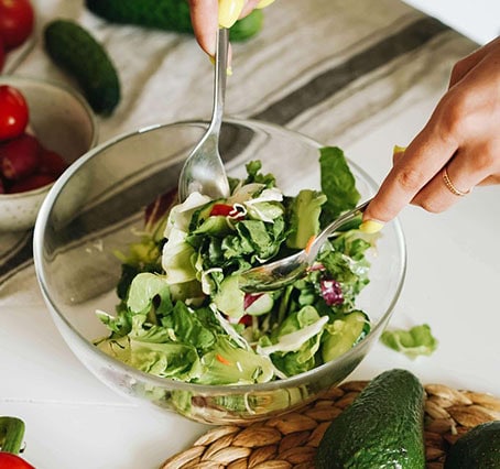 recipes Hands expertly mixing a fresh green salad in a glass bowl with two silver utensils—a true veganiac delight. The vibrant mix includes lettuce, tomatoes, and other greens, while avocados and extra tomatoes patiently await their turn on a light-colored tablecloth.