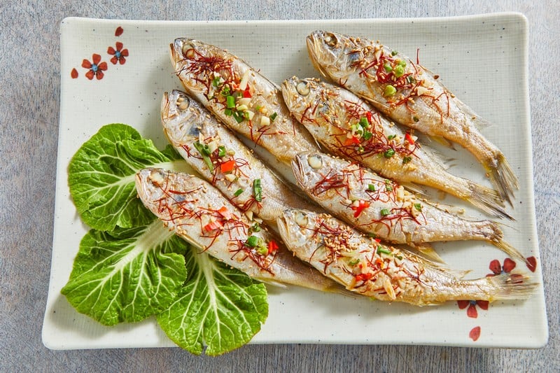 The image shows a beautifully arranged platter of steamed or grilled small fish, garnished with finely chopped green onions, red chili flakes, and shredded ginger. The fish are presented on a rectangular ceramic plate with floral patterns, accompanied by fresh green lettuce leaves as a decorative side. The vibrant garnishes add a pop of color, enhancing the dish's visual appeal.  