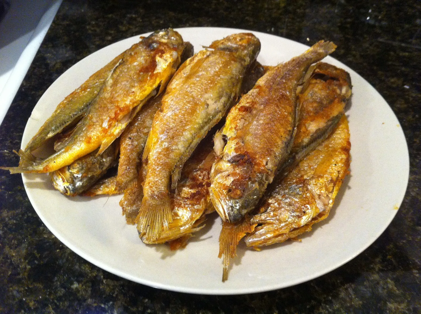 The image shows a plate of pan-fried small yellow croaker fish. The fish are golden brown with a crispy exterior, served simply on a white plate. Their texture suggests they have been lightly coated with flour or seasoning before frying, giving them a deliciously crunchy appearance.  