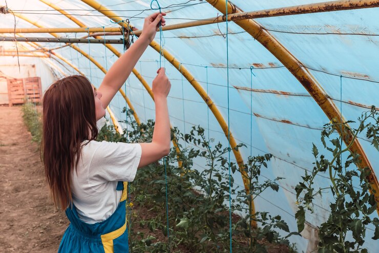 A close-up shot of a young female gardener working in a greenhouse, surrounded by lush green plants. She appears focused and dedicated, highlighting the nurturing aspect of gardening.