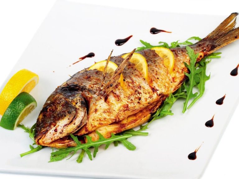 The image shows a beautifully plated grilled whole fish, likely a croaker fish, garnished with lemon slices, fresh arugula, and drizzles of a dark sauce, possibly balsamic glaze, on a clean white plate. The presentation is vibrant, with citrus wedges on the side adding a fresh touch.