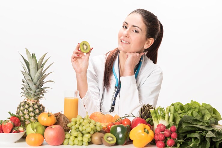 A woman in a white coat sits at a table filled with colorful fruits and vegetables, showcasing a healthy lifestyle.