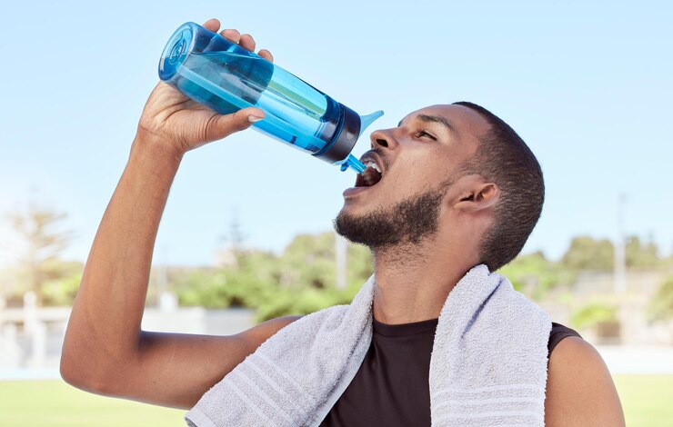 A man in athletic wear with a towel draped around his shoulders drinks water from a blue reusable bottle outdoors on a sunny day, with a blurred background of greenery and a clear blue sky.