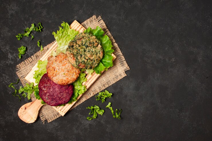 A wooden serving board with three colorful plant-based burger patties placed on fresh lettuce leaves, set on a dark textured background. The patties include a red beet-based patty, a green herb-infused patty, and a natural brown patty. Scattered lettuce pieces add a fresh and rustic touch to the presentation.