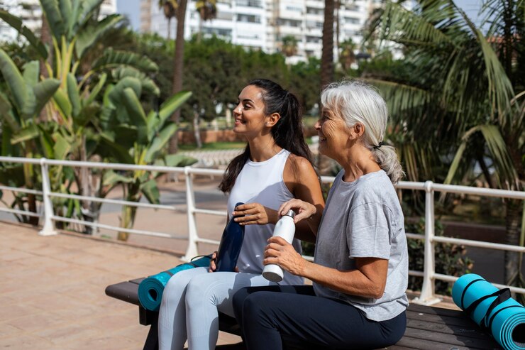 "An older woman and a younger woman sitting on an outdoor bench with yoga mats, enjoying a break and holding water bottles. They are dressed in activewear and smiling, with a tropical background featuring palm trees and greenery, indicating a relaxed and healthy lifestyle."