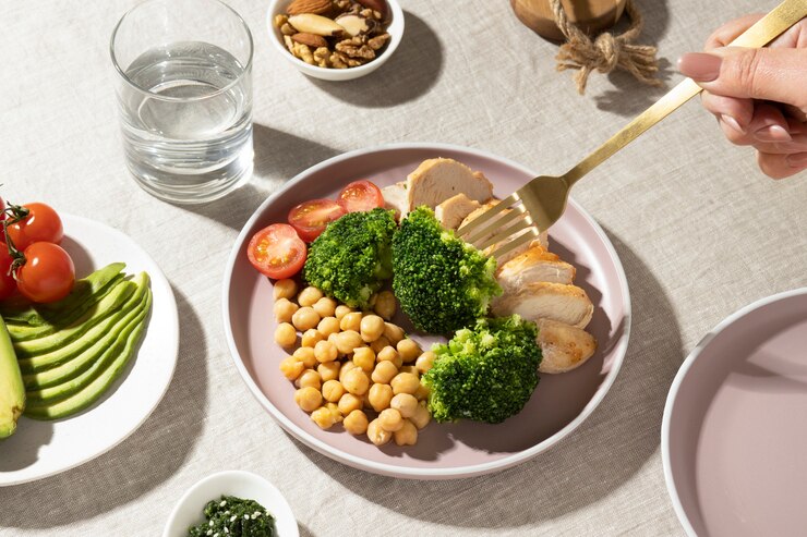 "A balanced meal with sliced grilled chicken breast, broccoli florets, cherry tomatoes, and chickpeas served on a light pink plate. Nearby are a plate of sliced avocado and cherry tomatoes, a glass of water, and small bowls with nuts and seeds. A person is holding a golden fork, ready to eat."