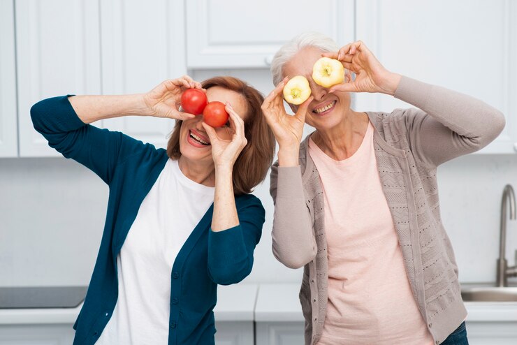 "Two joyful older women standing in a kitchen, playfully holding vegetables and fruits over their eyes like glasses. One woman holds red tomatoes, and the other holds yellow apples. They are smiling and enjoying a lighthearted moment, emphasizing health and vitality."