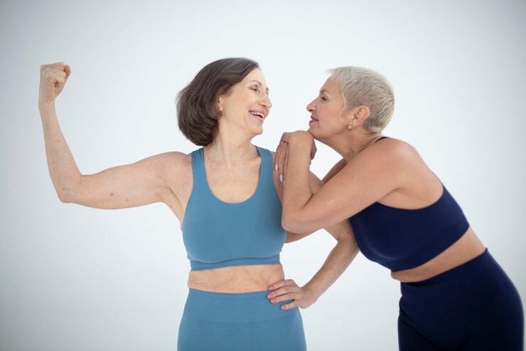 "Two confident older women in workout attire posing together in a studio. One woman flexes her arm, showcasing her strength, while the other leans on her arm with a smile. They are both wearing sports bras and leggings, celebrating fitness, strength, and friendship."