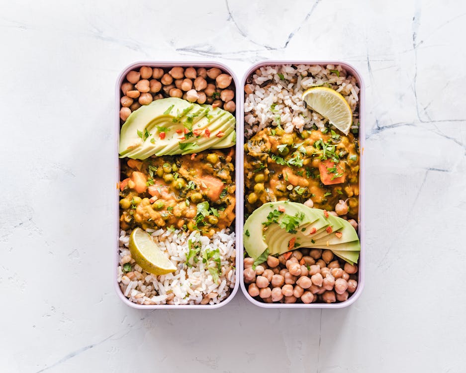 Two colorful lunch boxes filled with rice, beans, and sliced avocado, ready for a nutritious meal. The vibrant ingredients are neatly arranged.