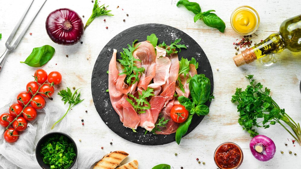 "A rustic food arrangement featuring a black slate plate with thinly sliced prosciutto, garnished with arugula, basil, and cherry tomatoes. Surrounding the plate are ingredients such as whole cherry tomatoes on the vine, fresh basil, parsley, a red onion, green herbs in a bowl, grilled bread slices, olive oil, mustard, and a small dish of sun-dried tomato paste, all set on a white wooden background."