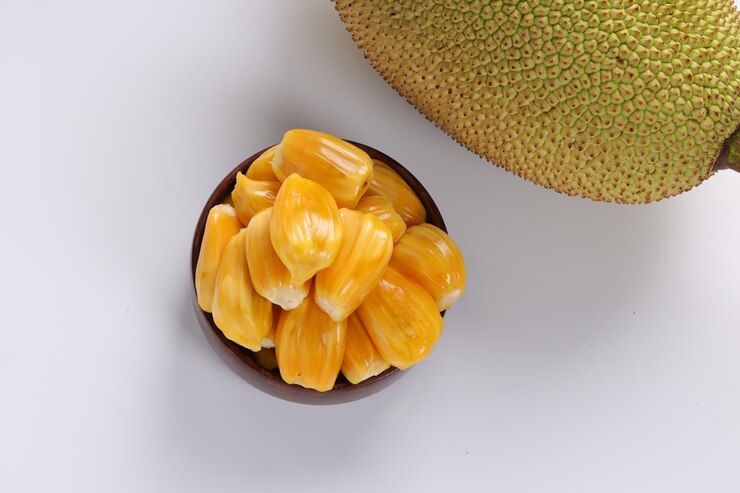 A beautifully arranged display of ripe jackfruit in a wooden bowl, set against a white textured background. The vibrant yellow color of the jackfruit contrasts with the neutral background, emphasizing its freshness and appeal.