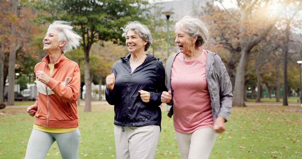 "Three joyful older women jogging together in a park on a cool day, wearing light jackets and activewear. They are smiling and enjoying each other's company, surrounded by trees with autumn leaves, symbolizing friendship, vitality, and an active lifestyle."