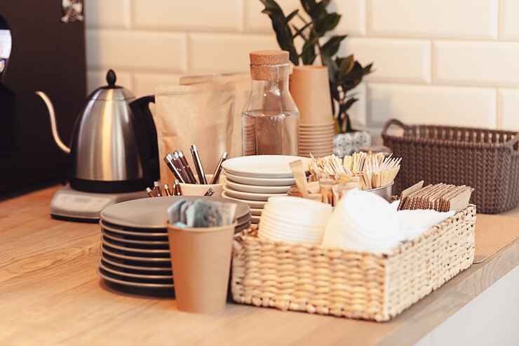 A cozy coffee station setup on a wooden countertop featuring stacked plates, cups, utensils, and a wicker basket holding disposable cups and stir sticks. A stainless steel electric kettle, a glass water bottle, and paper bags of coffee or tea are arranged neatly, with a small plant and woven storage basket adding a touch of decor in the background.