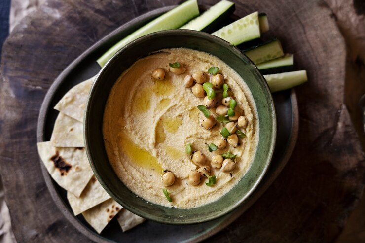 "Bowl of creamy hummus topped with whole chickpeas, a drizzle of olive oil, and fresh herbs, served on a plate with cucumber sticks and pita bread wedges. The presentation sits on a rustic wooden surface, highlighting a healthy Mediterranean snack."