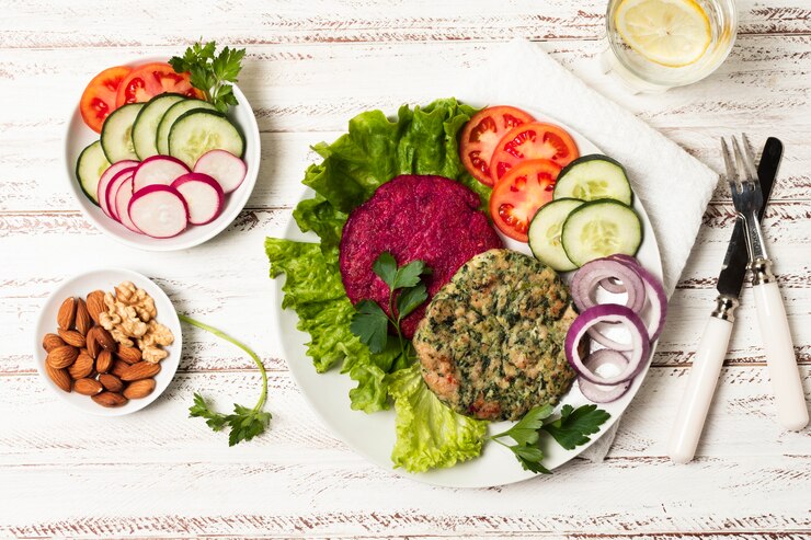 A colorful plant-based meal on a white wooden table featuring a plate with beet and herb-infused burger patties placed on lettuce leaves. The dish is garnished with fresh tomato slices, cucumber, red onion rings, and parsley. Nearby, small bowls hold a mix of almonds and walnuts, and sliced radishes and cucumbers. A glass of water with lemon and cutlery complete the elegant presentation.