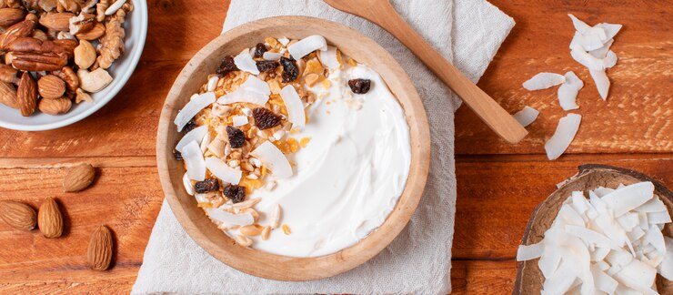 "Wooden bowl filled with yogurt topped with coconut flakes, raisins, nuts, and seeds, placed on a cloth napkin on a wooden table. Surrounding the bowl are additional nuts and coconut flakes, highlighting a healthy, wholesome breakfast option."