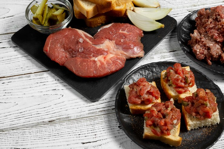 "A selection of ingredients and appetizers on a rustic white wooden table. Raw steak on a black slate board with sliced onions, bread slices, and a small bowl of pickles. Beside it, a black plate with toasted bread topped with diced tomatoes and herbs. There is also a portion of minced meat on a separate plate."