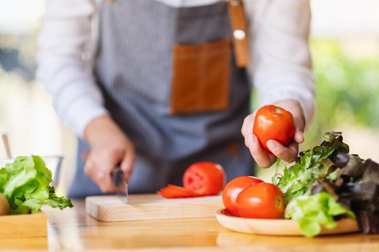 A person wearing a striped apron slicing a tomato on a wooden cutting board, with fresh lettuce and whole tomatoes on the counter in a bright kitchen setting.