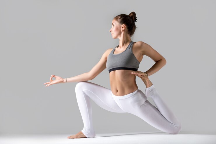 "Young woman in activewear performing a yoga pose on a gray background. She is in a low lunge position with one arm extended forward in a mudra and the other reaching back to hold her foot, displaying flexibility and balance."