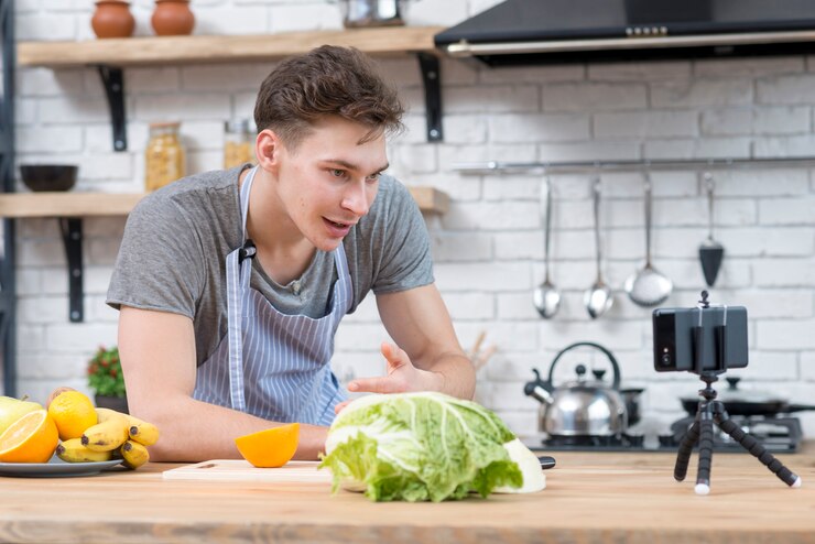 A man in an apron holds a camera while chopping vegetables, showcasing the plant-based cooking trend among vegan influencers.