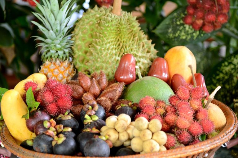 A vibrant assortment of tropical fruits displayed in a basket, featuring a durian, pineapple, bananas, and a variety of colorful fruits like rambutan, mangosteen, rose apples, and longans, surrounded by lush greenery.
