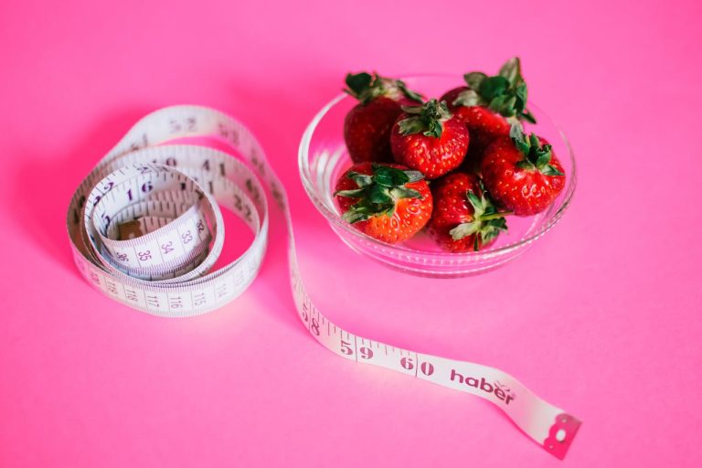 The image shows a glass bowl filled with fresh, vibrant strawberries placed on a bright pink background. Next to the bowl, a measuring tape is artistically coiled, symbolizing themes of healthy eating, dieting, or fitness.