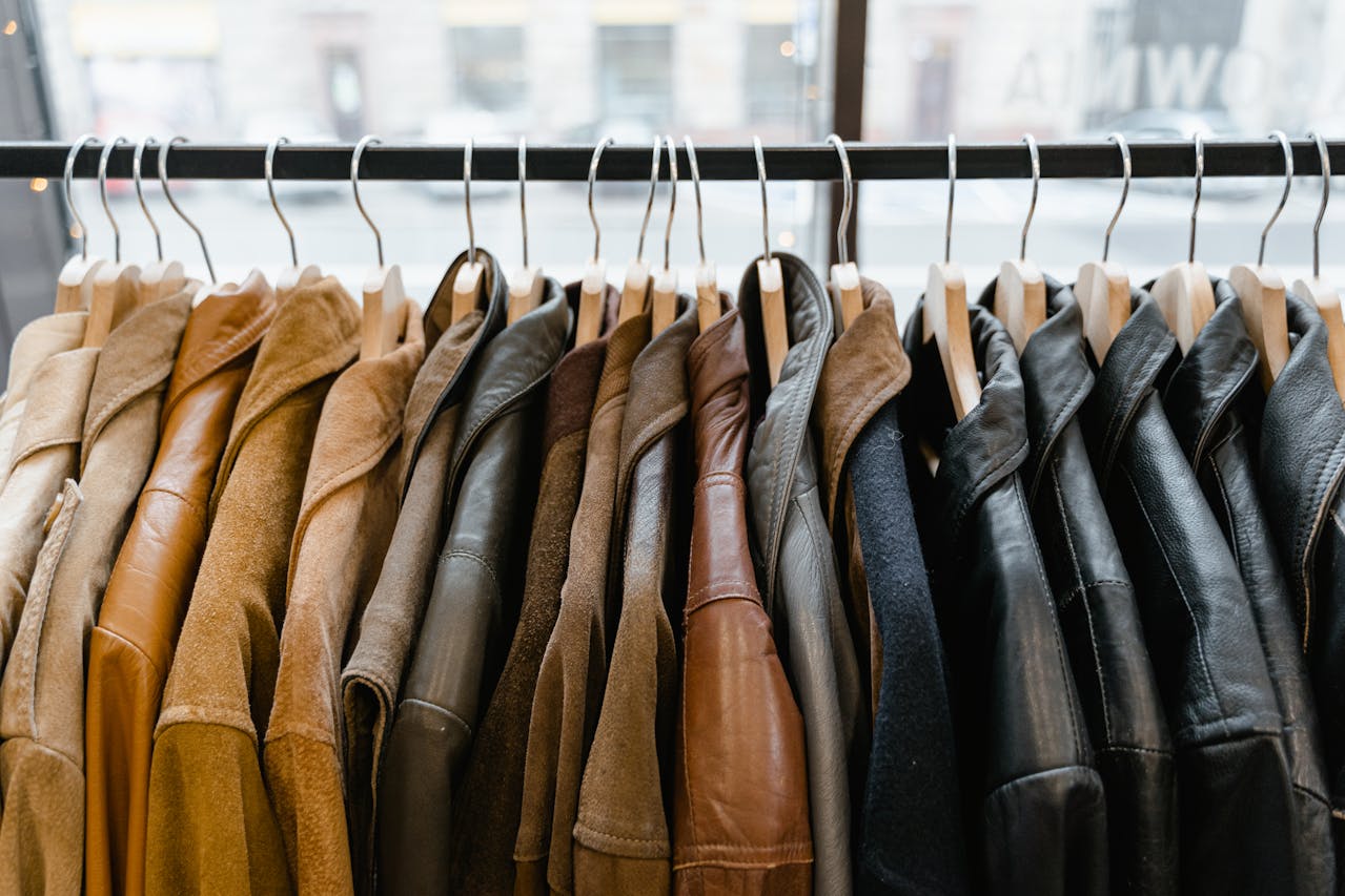 The image shows a rack of various jackets hanging on wooden hangers. The jackets come in different materials and colors, including brown, tan, and black, with a mix of leather and suede textures. The rack is placed indoors near a large window that overlooks a blurred urban street view.