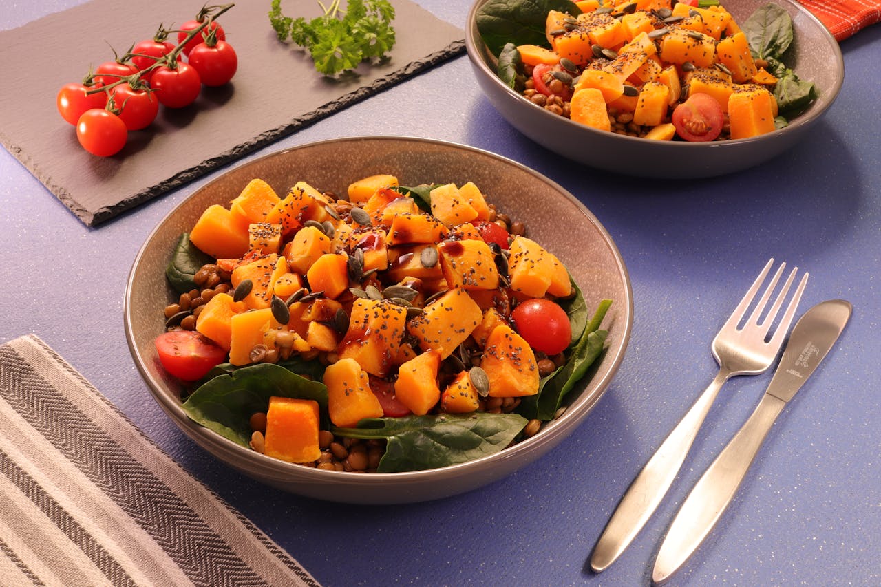The image features a vibrant salad bowl filled with roasted pumpkin chunks, cherry tomatoes, lentils, fresh spinach leaves, and topped with pumpkin seeds and a drizzle of dressing. The presentation is enhanced with a sprig of parsley and cherry tomatoes on a slate board in the background, alongside a fork and knife on the table, creating a fresh and appetizing setting.  