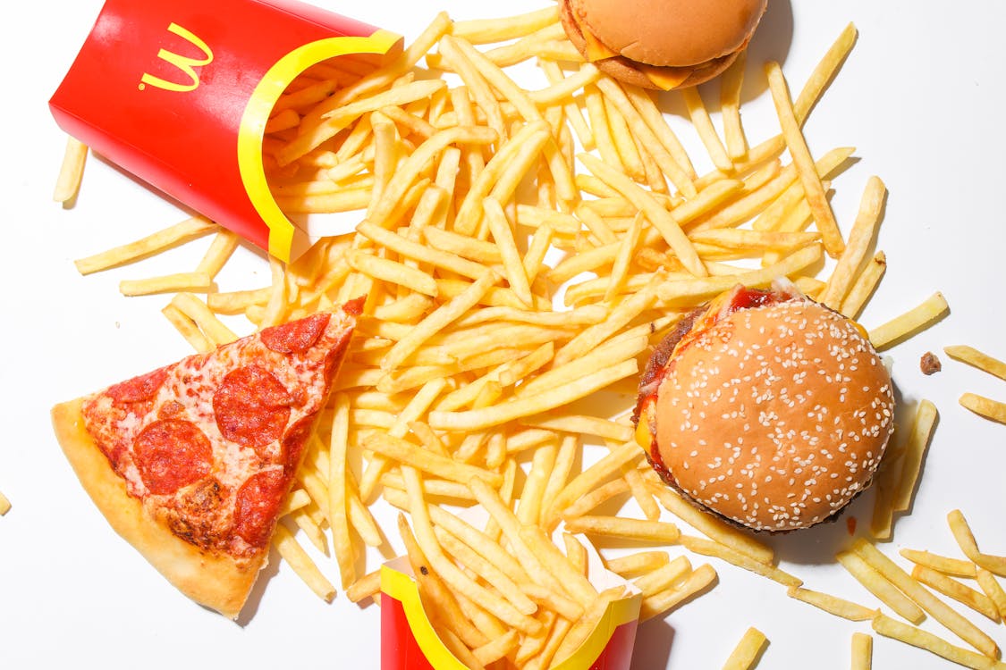 A gluten-free hamburger, fries, and pizza are beautifully arranged on a white surface, to avoid for a weight loss meal.