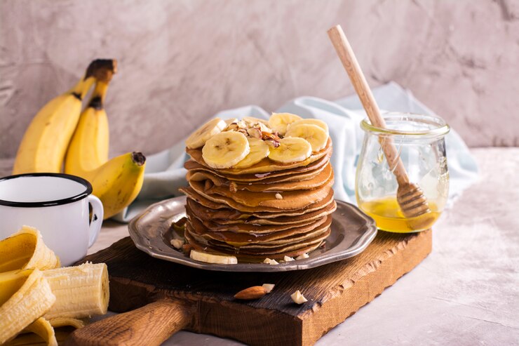 A wooden plate holds a stack of vegan banana oat pancakes topped with sliced bananas and nuts.