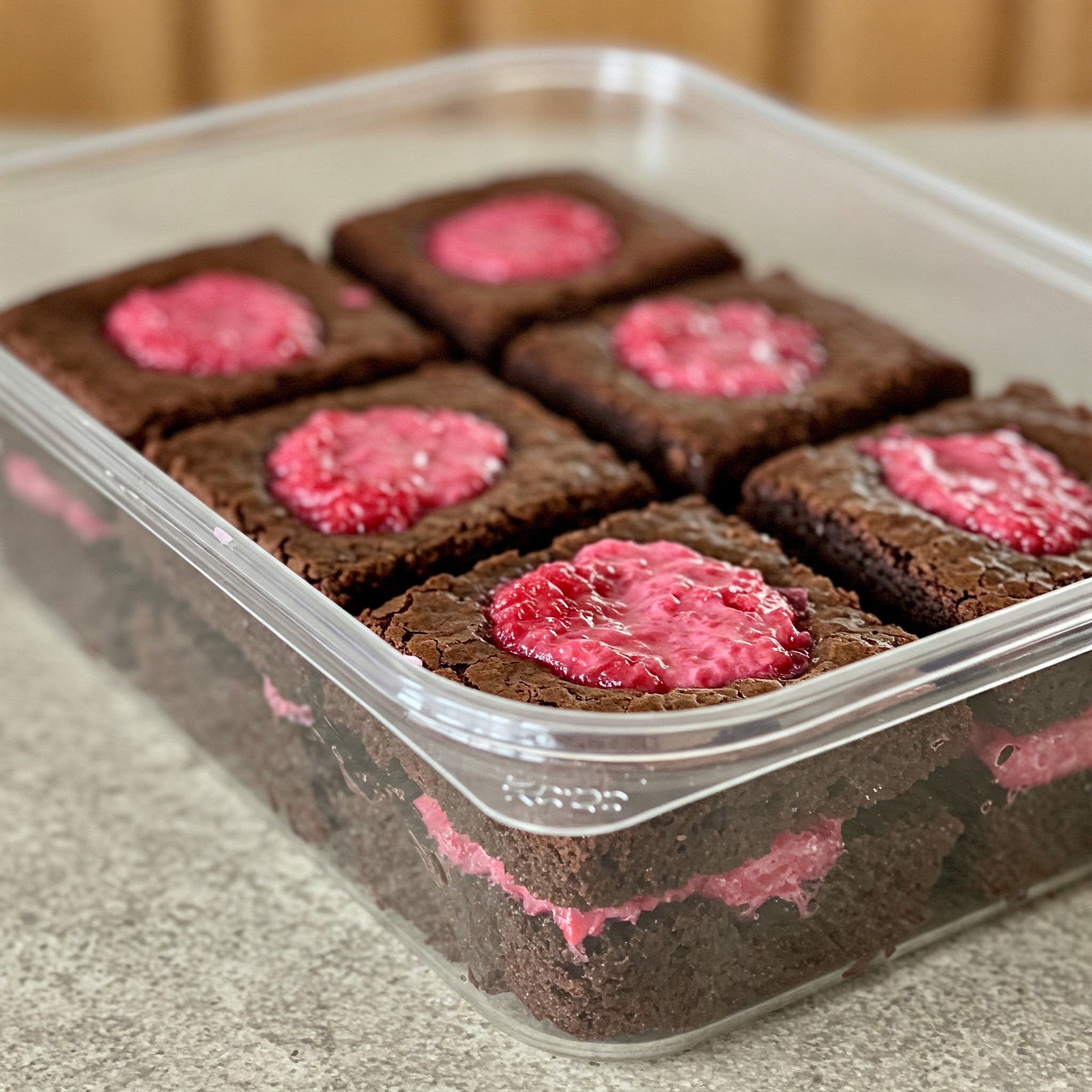 A plastic container holds six Valentine's Raspberry Truffle Brownies, showcasing their rich chocolate and raspberry layers.