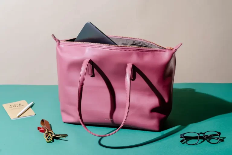 A stylish mauve tote bag with a structured design and two handles, sitting on a green surface. The bag is partially unzipped, revealing a laptop inside. Surrounding the bag are everyday essentials, including a pair of black-rimmed glasses, a set of keys with a leather keychain, a small notebook labeled 'Field Notes,' and a pen, all neatly arranged.