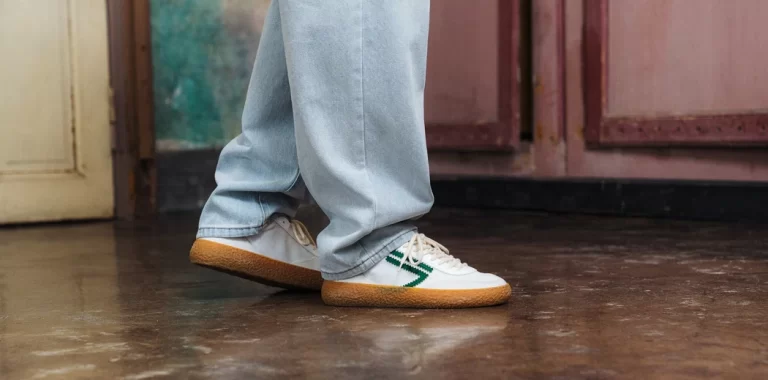 Top 10 Best Vegan Sneakers for 2025 A close-up of a person wearing white sneakers with green stripes and a gum sole, paired with loose-fitting light blue jeans. The individual is standing on a polished concrete floor with a vintage-style interior in the background, featuring aged pink and brown walls and a wooden door.