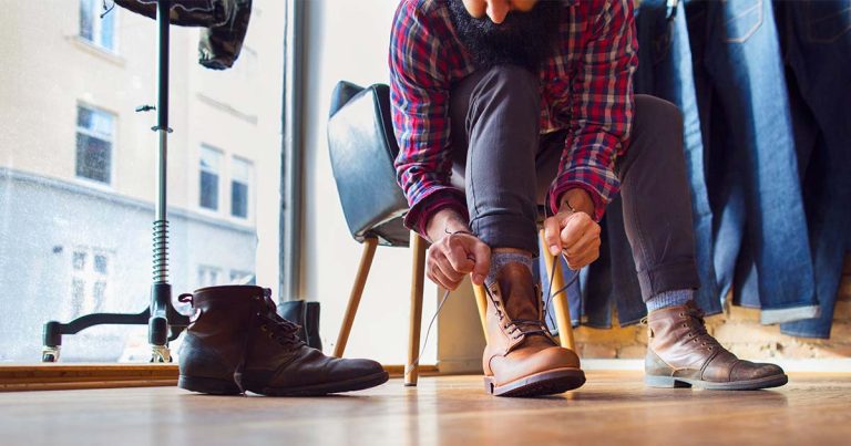 Top 10 Best Vegan Boots for 2025 A bearded man wearing a red plaid shirt and dark pants sits on a chair inside a clothing store, tying the laces of a new pair of brown leather boots. A worn black pair of boots is placed on the wooden floor beside him. The background features denim jeans hanging on a rack and a large window overlooking a street with buildings.