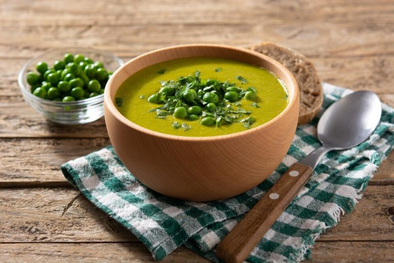 A bowl of fresh green pea and mint soup with peas, accompanied by bread, on a wooden table.