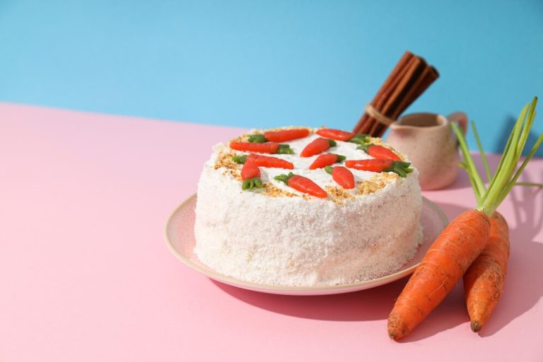 Vegan carrot cake topped with cream cheese frosting and decorative carrots, set against a pink background.