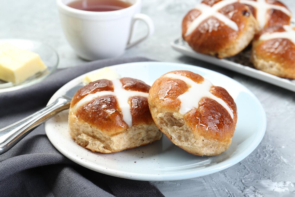 A plate of hot cross buns with butter, accompanied by a cup of tea, perfect for an easy vegan Easter brunch.