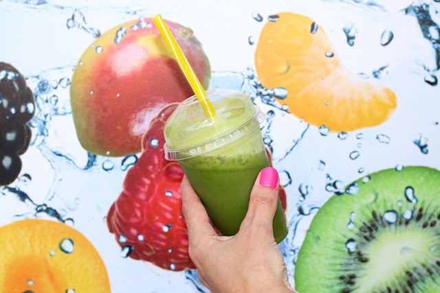 A person showcases a green smoothie in front of a fruit wall, featuring a pistachio smoothie recipe perfect for summer.
