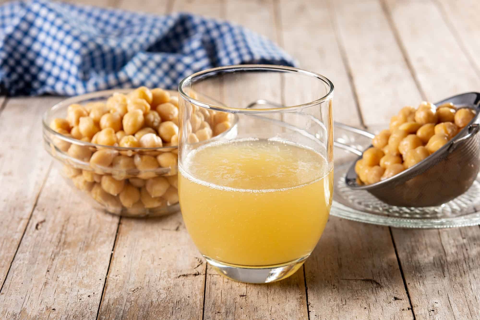 A glass of aquafaba, the liquid from cooked chickpeas, with bowls of chickpeas in the background on a rustic wooden table.