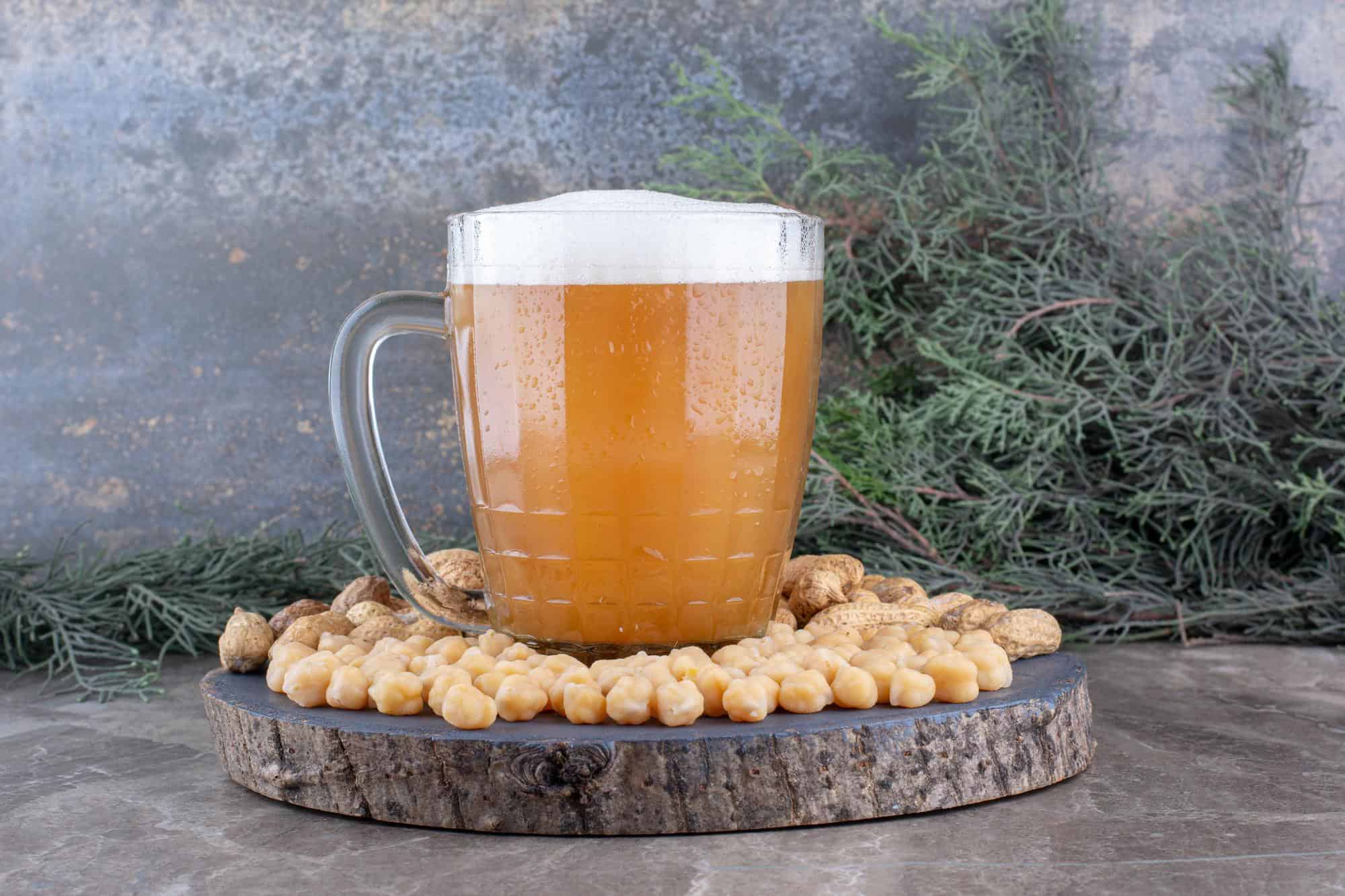 A clear glass mug of vegan Butterbeer made with aquafaba foam, surrounded by chickpeas on a wooden display, with greenery in the background.