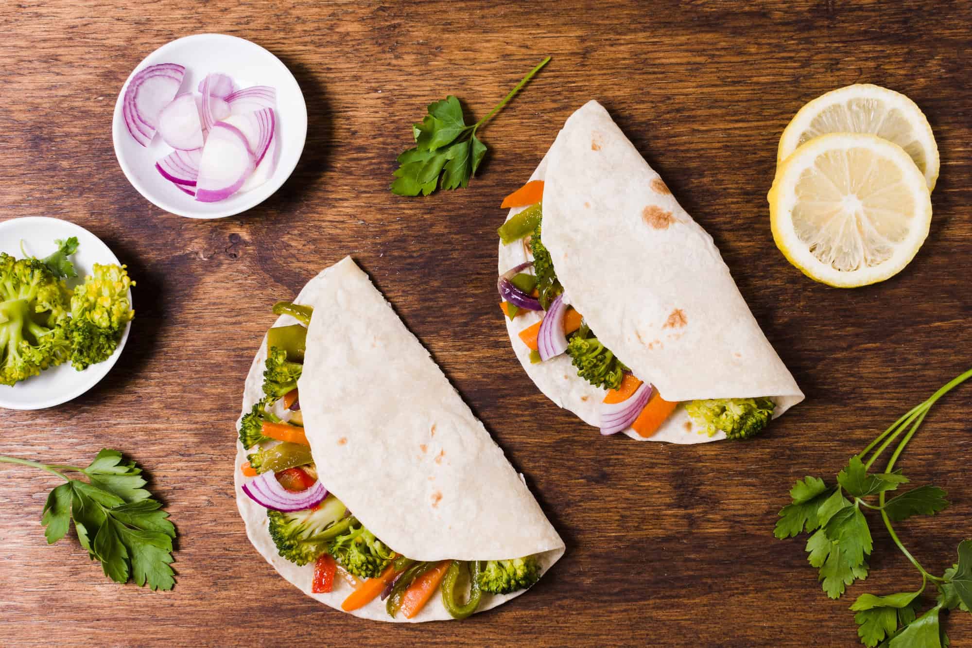 Top view of vegan vegetable tortilla wraps filled with broccoli, peppers, and onions on a wooden table with parsley and lemon slices
